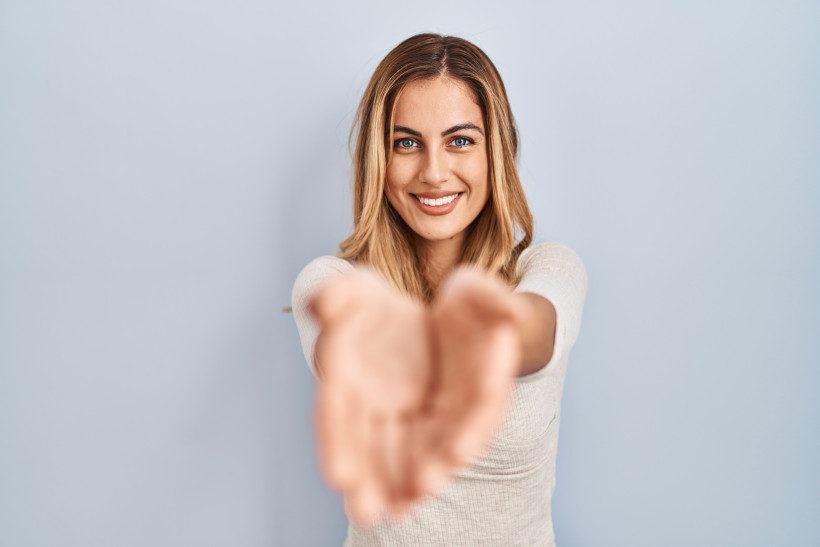 young blonde woman standing isolated background smiling with hands palms together receiving giving gesture hold protection